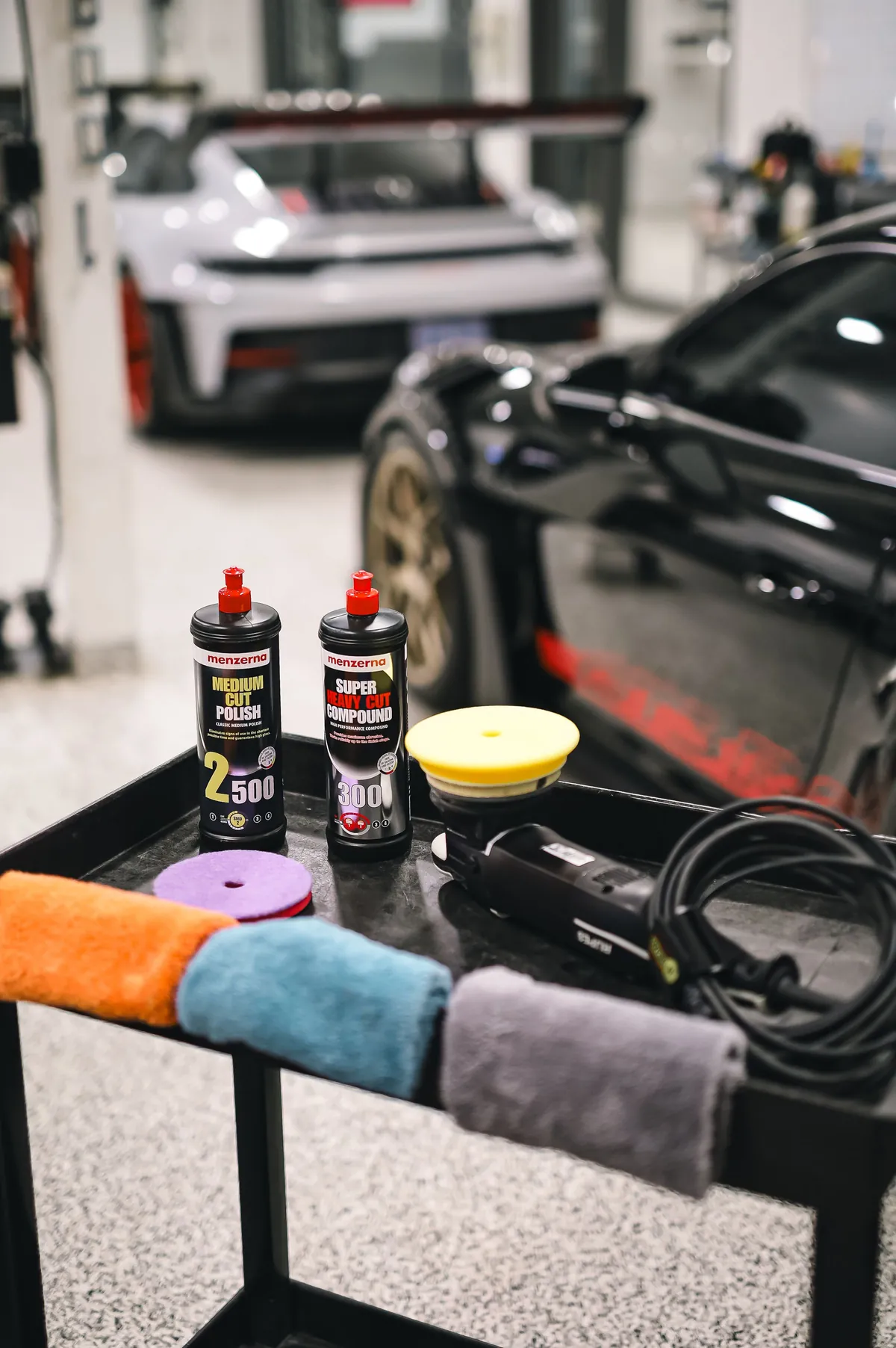 Polishes, pads and towels on a cart beside a sports car