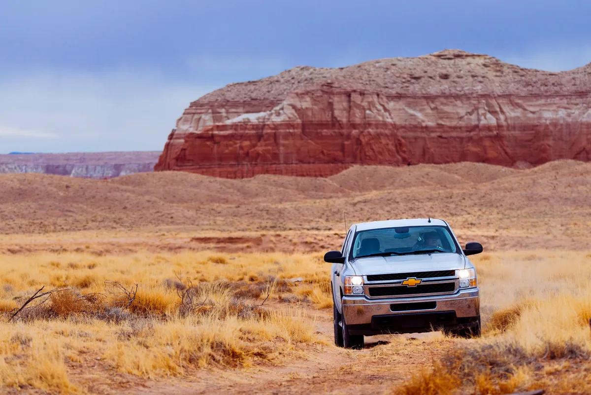 A silver pickup on a dirt road in front of a red rock mesa